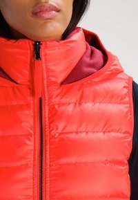Gilet imbottito rosso con colletto alto e chiusura con zip, caratterizzato da un design trapuntato e una texture lucida, sovrapposto a un indumento scuro.