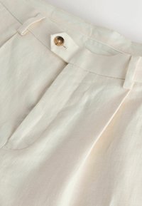 Nærbillede af en cremefarvet bukselinning med en enkelt brun knap og bæltestropper, der viser stoftekstur og syningsdetaljer.