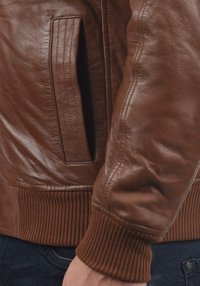 Närbild av en person som bär en brun läderjacka med ruggade ärmslutar och midja, samt mörkblå denimjeans.