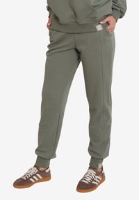 Oliwkowe dresy z elastycznymi mankietami, bocznymi kieszeniami i naszywką z logo. Wykonane z miękkiego, teksturowanego materiału zapewniającego komfort.