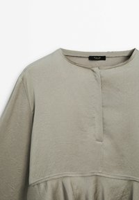Haut gris clair avec un col rond, une patte de boutonnage discrète à l'avant, des manches longues, et une texture lisse. Comprend un drapé de tissu au niveau de l'ourlet.