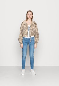 Jaqueta de camuflagem bege, cortada, com capuz e punhos elásticos, combinada com jeans skinny azuis claros e ténis brancos.