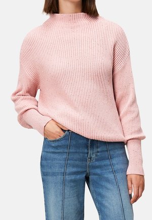 Vrouw draagt een roze geribbelde trui met lange mouwen en blauwe high-waisted jeans met zichtbare voorste naden.