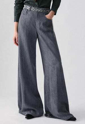 Personne portant un pantalon gris à jambes larges avec une ceinture chaîne argentée et des chaussures noires pointues, main dans la poche droite sur fond uni.