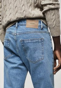 Jeans de mezclilla azul claro con corte de pierna recta. Presentan dos bolsillos traseros y un parche de cuero marrón con la etiqueta "MNG DENIM". Combinados con un suéter de punto trenzado.