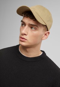 Cap features a beige, structured design with a curved brim. The individual wears a black, ribbed sweater with a crew neckline.