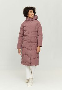 Langer, gepolsterter Mantel in Mauve, mit hohem Kragen und seitlichen Taschen, im gesteppten Design und mit Reißverschluss. Getragen über weißen Hosen und Sneakers.