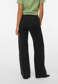 Personne portant un jean noir à jambes larges et une chemise verte à manches courtes, vue de dos sur un fond uni.