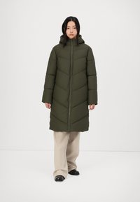 Abrigo acolchado verde oliva con cierre de cremallera, capucha y patrón en forma de chevron; combinado con pantalones de pierna ancha beige y zapatos negros.