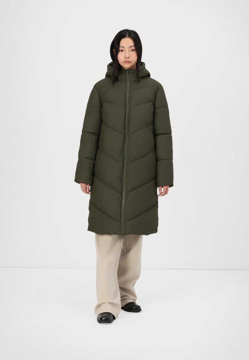 Abrigo acolchado verde oliva con cierre de cremallera, capucha y patrón en forma de chevron; combinado con pantalones de pierna ancha beige y zapatos negros.