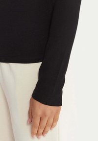 Camicia nera a maniche lunghe con design aderente, texture morbida e polsini a costine, abbinata a pantaloni di colore chiaro. L'immagine mostra un primo piano della manica.