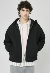 Haut à capuche noir avec une grande capuche, des poches avant et des poignets côtelés, superposé sur une chemise rose clair et une chemise à rayures.