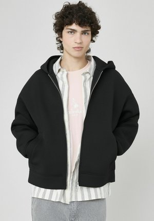 Haut à capuche noir avec une grande capuche, des poches avant et des poignets côtelés, superposé sur une chemise rose clair et une chemise à rayures.