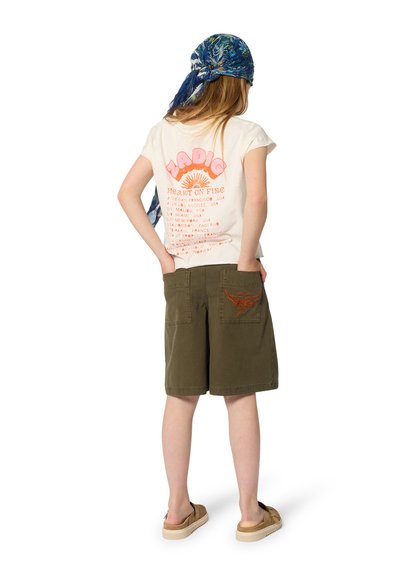 Enfant portant un foulard bleu à fleurs, un t-shirt blanc à motif graphique, un short vert olive avec un motif brodé rouge, et des chaussures slip-on beige, vu de dos.