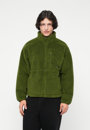 Giovane con capelli scuri che indossa una giacca in pile verde con cerniera e pantaloni neri, davanti a uno sfondo bianco semplice.