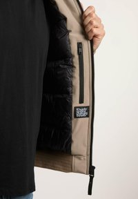 Beige ytterjakke med svart, vattert fôr, med sidelomme med glidelås og etikett med "Khujo"-merke. Strukturert stoff ved kanten.