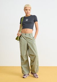Svart kort t-shirt med vit stjärnlogga, oliva gröna lösa byxor och mönstrade sneakers, mot en ljus bakgrund.