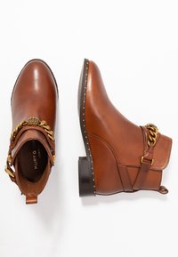 Botas de cuero marrón hasta el tobillo con punta redonda, decoración de cadena dorada y detalles de tachuelas a lo largo de la suela. Incluyen una hebilla lateral y una tira de agarre.
