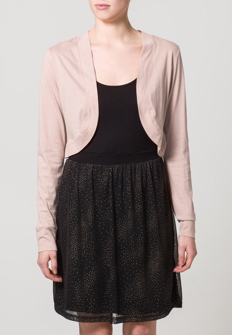 Ljusrosa kortärmad cardigan med långa ärmar, stylad över en svart ärmlös topp och en svart kjol med guldprickar.