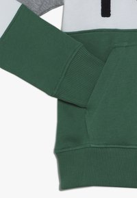 Felpa verde con polsini e orlo a costina, caratterizzata da un design superiore grigio e bianco e una tasca a marsupio. Tessuto morbido.