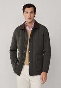 Donkergroene gewatteerde jas met een bruine corduroy kraag, sluiting met drukknopen aan de voorkant, twee zijzakken, daarover een beige trui en een gestreept shirt.