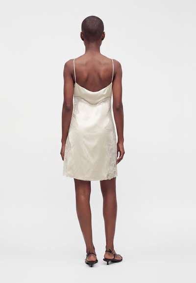 Femme aux cheveux courts vêtue d'une robe en satin ivoire sans manches avec des détails en dentelle et des sandales noires à talons bas, debout, tournée dos à l'avant.