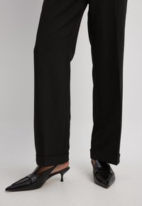 Schwarze Slingback-Absätze mit spitzem Zehenbereich, Obermaterial aus strukturiertem Leder und kleinem konisch zulaufendem Absatz, kombiniert mit weiten schwarzen Hosen, die leicht gekrempelt sind.