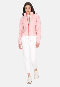 Lyserød quiltet jakke med høj krave og kontrastlynlås, parret med hvide sweatpants. Modellen bærer sneakers i lyse farver og med accenter.