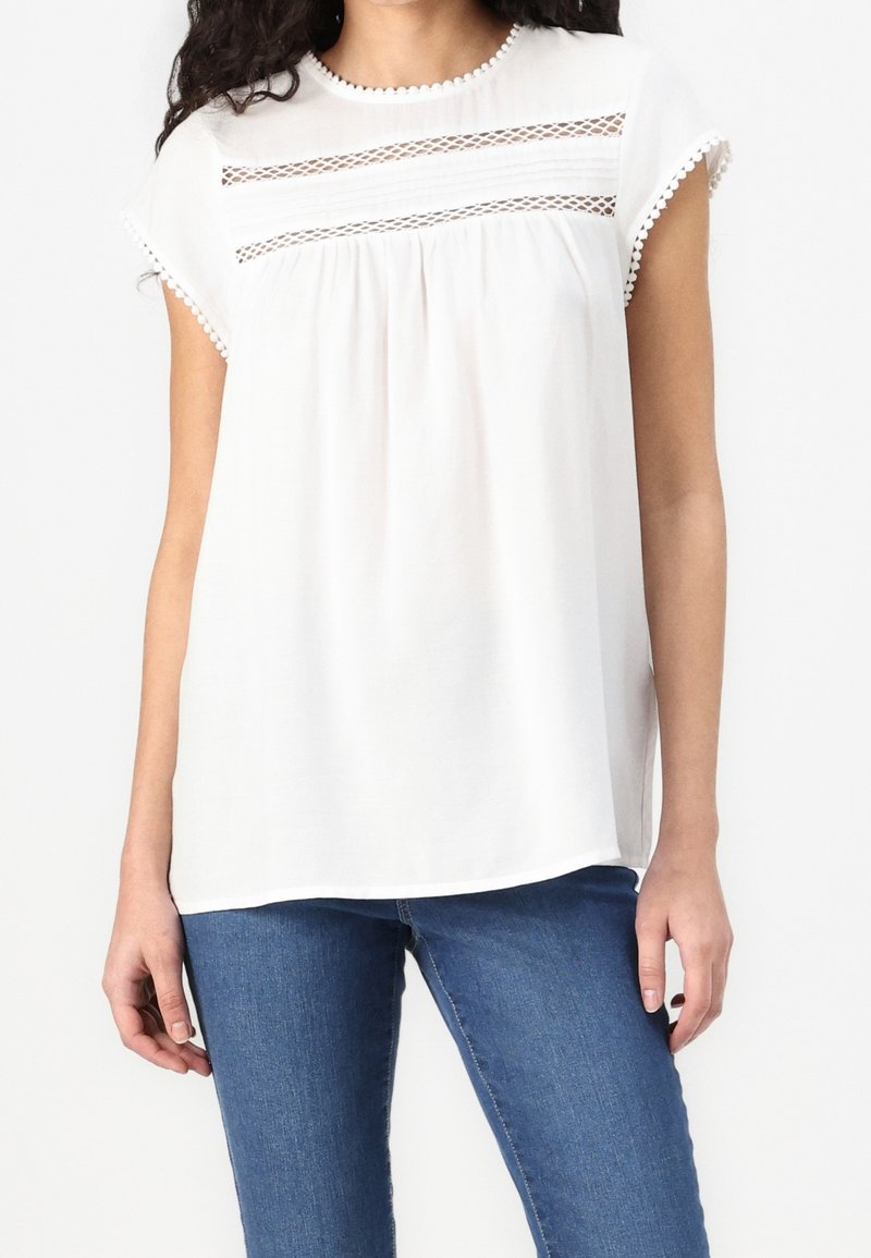 Femme portant un chemisier blanc à manches courtes avec des détails en dentelle et un jean bleu, debout devant un fond clair uni.