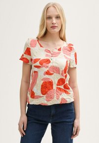 T-shirt à manches courtes en crème avec des motifs abstraits rouges et roses, présentant un col rond, une patte de boutonnage à trois boutons et une texture froissée.