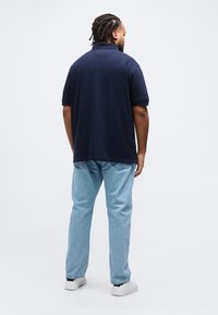 Άνδρας με πλεξούδες, φορώντας μπλε πόλο πουκάμισο, ανοιχτόχρωμα τζιν και λευκά αθλητικά παπούτσια, στέκεται με την πλάτη γυρισμένη σε απλό λευκό φόντο.