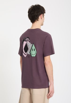 Homme portant un t-shirt violet avec des motifs géométriques abstraits et des visages souriants au dos, assorti à un short beige, vu de dos.