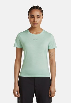 Frau mit geflochtenem Haar, die ein hellgrünes T-Shirt mit kurzen Ärmeln und schwarze Hose trägt, vor einem schlichten weißen Hintergrund stehend.