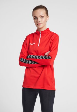 Rode sportieve top met lange mouwen, staande kraag, ritssluiting en een textuur op het lichaam. Zwart chevronpatroon op de mouwen. Merkenlogo zichtbaar.