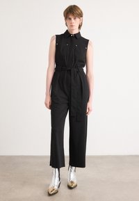 Mono negro sin mangas con cuello, bolsillos frontales y un cinturón en la cintura; tejido suave y piernas anchas, llevado con botas metálicas hasta el tobillo.