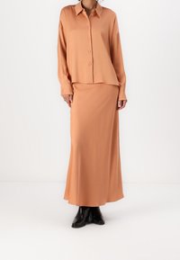 Camisa de corte suelto con botones y falda larga a juego en suave tela melocotón. Textura suave, cuello ancho, mangas largas y largo hasta los tobillos. Botas negras.