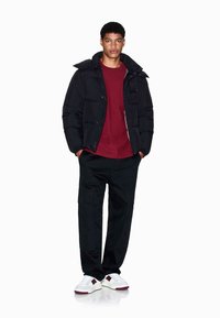 Schwarze Daunenjacke mit hohem Kragen, burgunderrohem Hemd, schwarzen Cargo-Hosen und weißen Turnschuhen mit roten Akzenten. Glatte Textur, lässiger Stil.