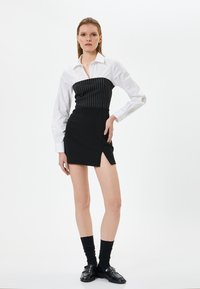 Vestito nero con un corsetto a righe, camicia bianca con colletto e uno spacco alto nella gonna. Abbinato a calze nere e mocassini.