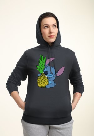 Person trägt einen dunklen Kapuzenpullover mit einem cartoonartigen Motiv eines Charakters, der eine große Ananas umarmt, die vorne aufgedruckt ist, hat die Hände in den Taschen und schaut nach oben.