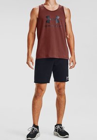 Czerwony bezrękawnik sportowy z czarnym logo, zestawiony z czarnymi szortami z białym logo, noszonymi z czarnymi butami sportowymi.