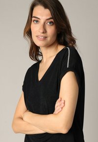 Haut noir à manches courtes avec un design en V, présentant un tissu texturé et des accents argentés sur les coutures des épaules.