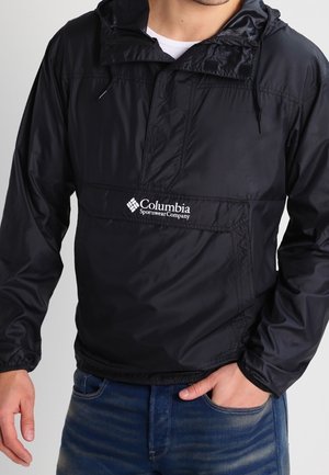 Mann trägt eine schwarze Columbia Sportswear Company Windjacke mit Kapuze über einem weißen Hemd, Hand in der Tasche der blauen Jeans.