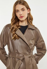 Ruskea nahkainen kaksirivinen trench-coat, jossa on suuri kaulus, vyötärölle sidottava vyö ja teksturoitu pinta. Sisältää sivutaskut ja nappikoristeet.