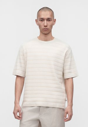 Homme à tête rasée portant une chemise à manches courtes texturée beige clair et un pantalon beige clair assorti, debout devant un fond blanc uni.