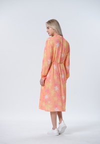 Langes, florales Kleid in Pfirsich mit pinken und gelben Blättern. Verfügt über lange Ärmel, eine taillierte Taille und weiße Sneakers.