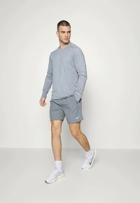 Mann som går, iført grå langermet skjorte, grå shorts, hvite sokker og hvite sneakers mot en enkel lys bakgrunn.