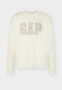 Krämfärgad tröja med långa ärmar, prydd med en stor "GAP"-logga i beige över bröstet. Ribbad krage och fåll. Mjuk textur.