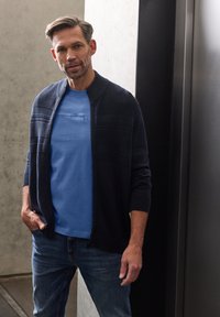 Modrý námořnický cardigan na zip s texturovaným vzorem přes světle modré tričko s kulatým výstřihem, doplněný džínami. Hladká látka a ležérní střih.