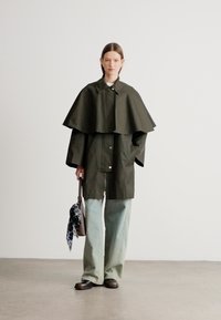 Casaco verde-oliva, largo e leve, com design estilo capa, gola grande e botões de pressão frontais. Combinado com calças largas azul claro.