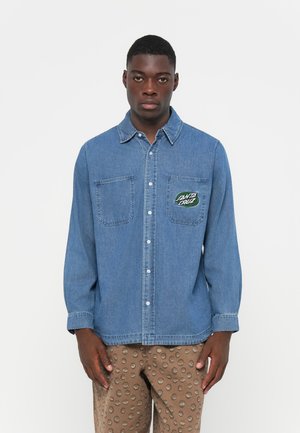lichtblauwe denim overhemd met knoopsluiting, twee borstzakken en een cirkelsgewijze groene geborduurde logo op de linkerzak. Lange mouwen.